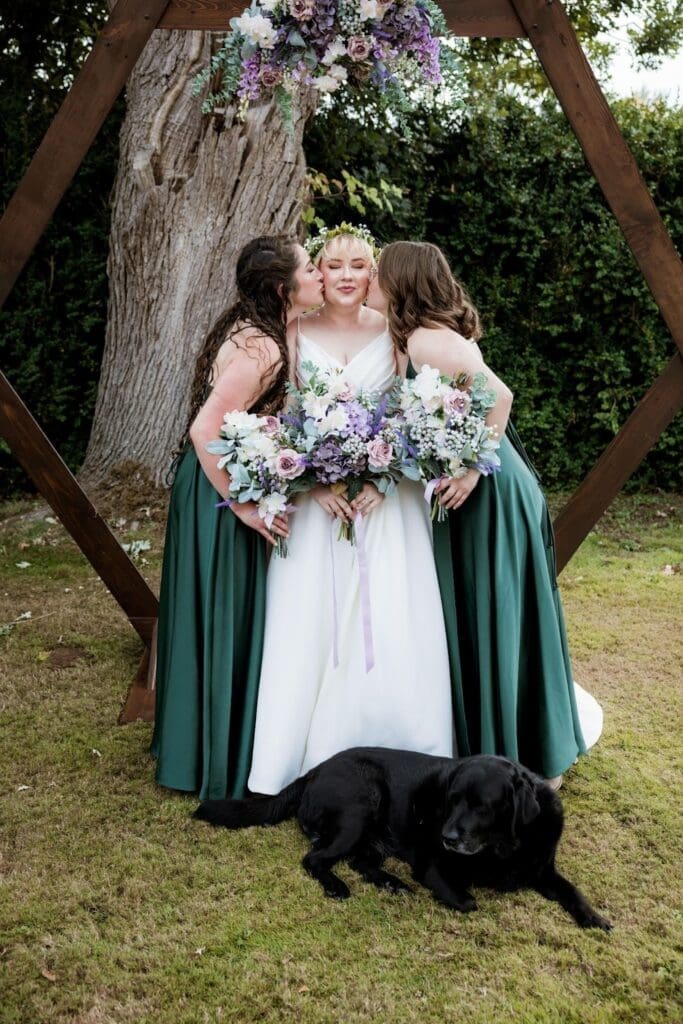Bridesmaids kissing Mandy on the cheeks under the arch in an outdoor barn wedding venue